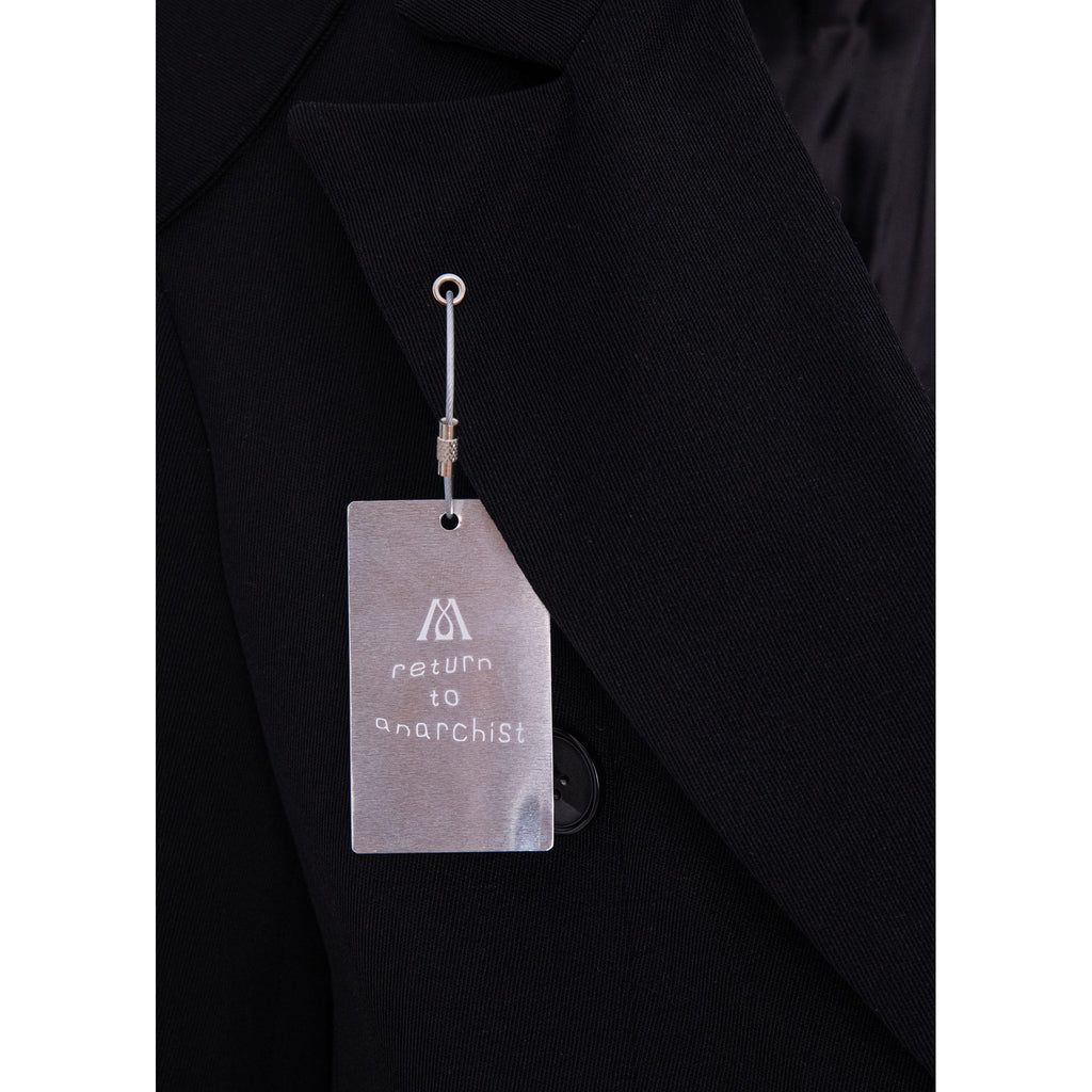Close-up of a black blazer lapel with a metal hangtag attached. The tag has a white logo and the text 'return to anarchist' printed on it.