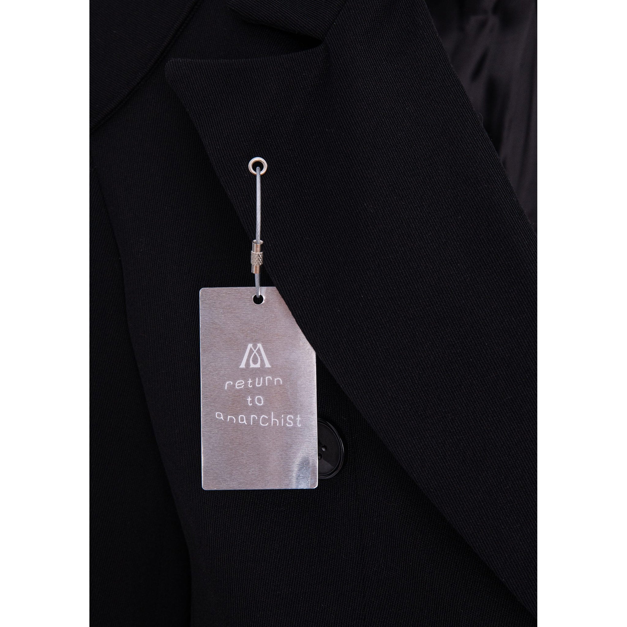 Close-up of a black blazer lapel with a metal hangtag attached. The tag has a white logo and the text 'return to anarchist' printed on it.